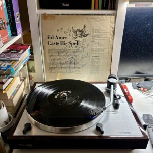 Ed Ames - my cup runneth over - Vinyl B - Ein Thorens Plattenspieler mit einem Albumcover von Ed Ames "Casts His Spell" obenauf, das die schwarze Vinyl-Schallplatte zeigt. Der Plattenspieler befindet sich auf einem Holzschrank, der seine silberne Oberfläche zeigt. Das Albumcover zeigt eine Zeichnung eines Cherubs und den Titel in einem Vintage-Design. Bücher und andere Gegenstände befinden sich im Hintergrund auf Regalen.