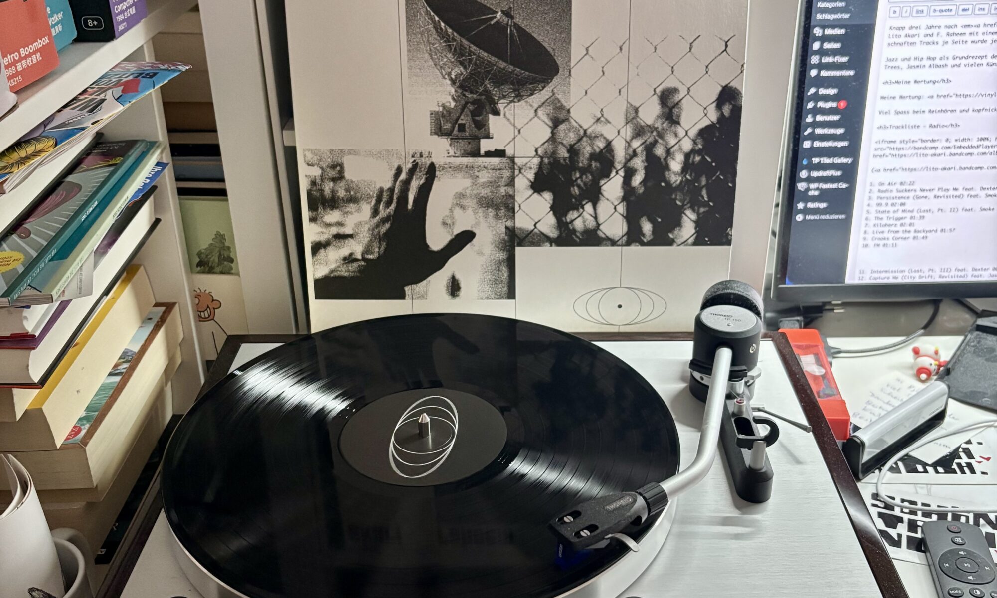 Akari raheem radio vinyl a - Eine Schallplatte dreht sich auf einem Thorens-Plattenspieler aus Silber und dunklem Holz, im Hintergrund ist das Albumcover für "akari raheem radio" zu sehen. Die Albumcover-Gestaltung zeigt eine Collage mit einer Satellitenschüssel, einer Silhouette einer Hand vor einem Maschendrahtzaun und verschwommenen Figuren. Rechts neben dem Albumcover befindet sich ein Computermonitor, der Website-Inhalte anzeigt.