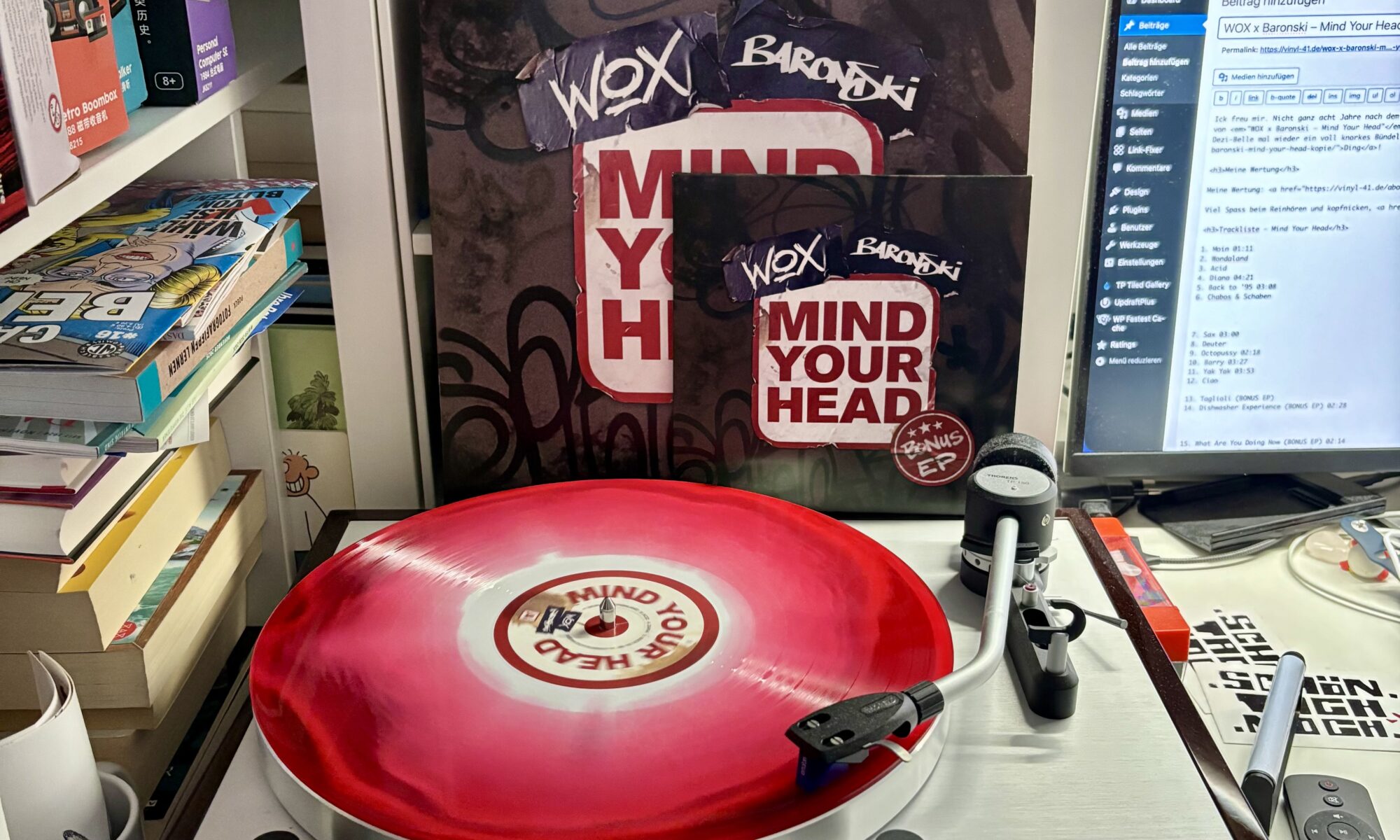 Wox x baronski mind your head vinyl - Ein Thorens-Plattenspieler mit einer rot-weißen Vinylplatte, die abgespielt wird, mit der Aufschrift "MIND YOUR HEAD" auf dem Mittellabel, ist im Fokus. Regale mit Büchern und Albumcovern befinden sich im Hintergrund, zusammen mit einem Computerbildschirm, der eine Website und Text anzeigt, einschließlich der Namen "WOX" und "Baronski".