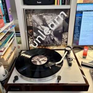 2econd class citizen unlearn vinyl a - Ein Plattenspieler mit einer schwarzen Vinyl-Schallplatte, die sich dreht, einem Schallplatten-Cover mit abstrakter Schwarz-Weiß-Kunst und dem Wort "unlearn" in Weiß aufgedruckt, und einem Computermonitor, auf dem eine Webseite angezeigt wird.