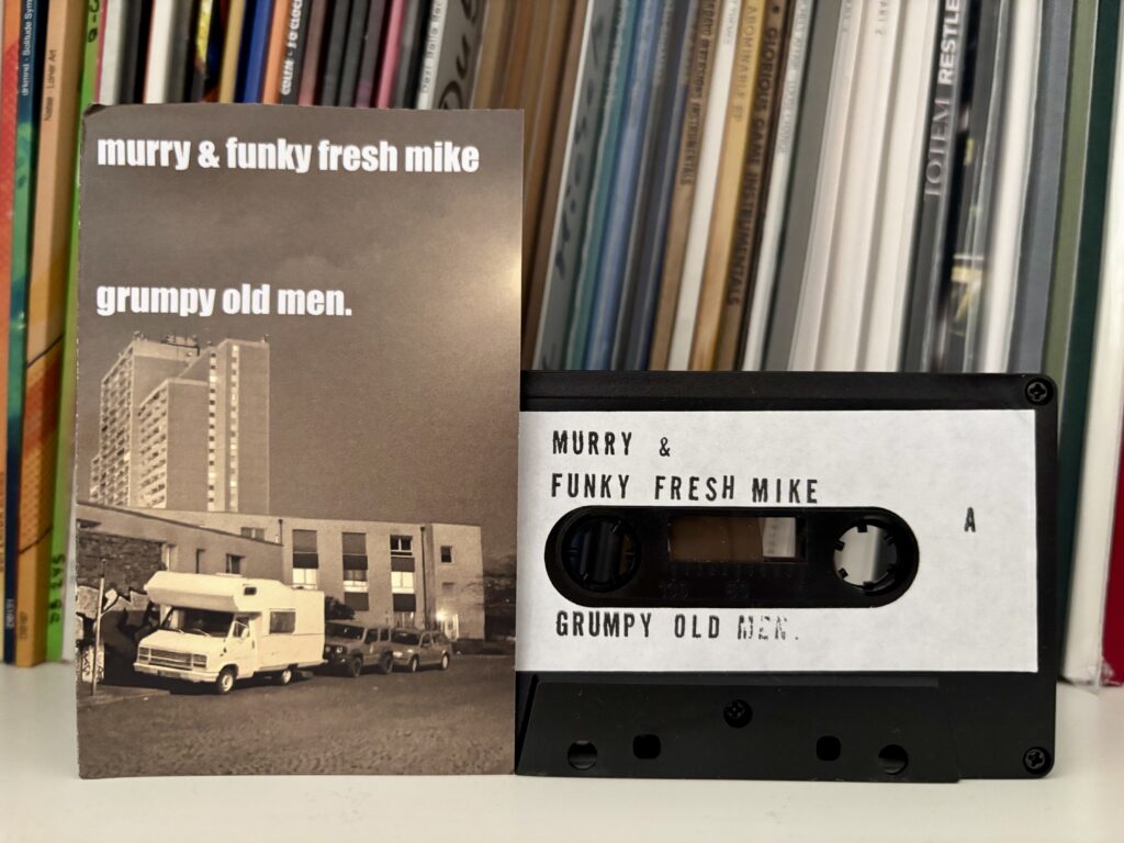 Murry funky fresh mike grumpy old men tape - Eine Kassette mit der Beschriftung „murry & funky fresh mike grumpy old men“ liegt neben ihrer passenden J-Card, die ein Schwarz-Weiß-Bild eines weißen Wohnmobils zeigt, das vor Apartmentgebäuden unter einem dunklen Himmel geparkt ist.