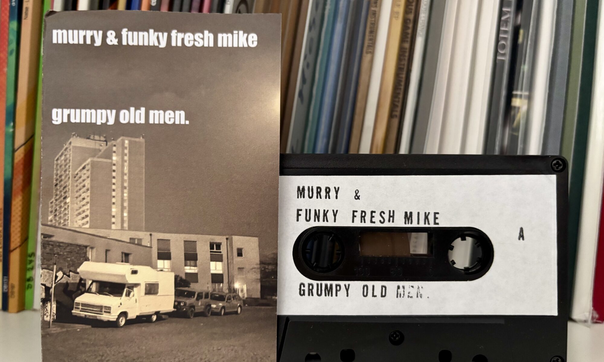 Murry funky fresh mike grumpy old men tape - Eine Kassette mit der Beschriftung „murry & funky fresh mike grumpy old men“ liegt neben ihrer passenden J-Card, die ein Schwarz-Weiß-Bild eines weißen Wohnmobils zeigt, das vor Apartmentgebäuden unter einem dunklen Himmel geparkt ist.