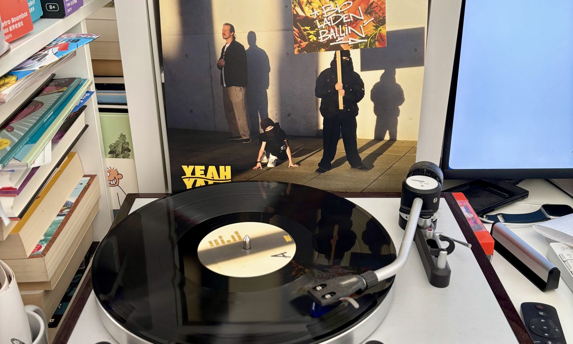 Rapper henning bioladen ballin yeah yall vinyl a - Ein Vinyl-Schallplatte spielt auf einem Thorens-Plattenspieler, der vor einem Albumcover steht, das zwei Figuren und ein Banner mit der Aufschrift "BIOLADEN BALLIN" zeigt. Ein Computermonitor ist rechts sichtbar, und links sind Regale voller Bücher und anderer Gegenstände.