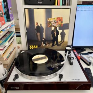 Rapper henning bioladen ballin yeah yall vinyl a - Ein Vinyl-Schallplatte spielt auf einem Thorens-Plattenspieler, der vor einem Albumcover steht, das zwei Figuren und ein Banner mit der Aufschrift "BIOLADEN BALLIN" zeigt. Ein Computermonitor ist rechts sichtbar, und links sind Regale voller Bücher und anderer Gegenstände.