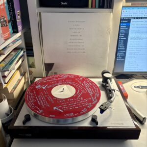 Flea honora nonesuch - seite d - Ein Plattenspieler mit einer roten Schallplatte. Die Platte hat handschriftliche Songtexte in Weiß. Im Hintergrund befindet sich ein Platten-Cover mit einer Trackliste, neben einem Computermonitor, der eine Webseite anzeigt.